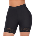 A close-up studio shot of a person's midsection wearing tight, high-waisted shapewear shorts in solid black. The shorts feature a smooth, compressive fabric body, a wide supportive waistband, and are finished with matching black floral lace trim at the hem of each thigh.